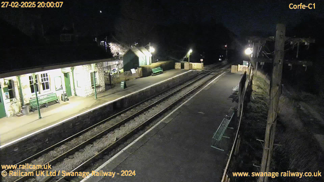 A dark night scene at a train station. The image shows an empty platform with several green benches and a stone building. There are two light sources illuminating the area, casting shadows. The railway tracks run along the platform, leading into the darkness. A wooden fence at the far end marks an exit. The time and date are displayed at the top left corner: 27 February 2025, 20:00:07.