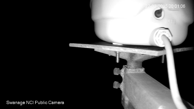 The image shows a close-up view of a white cylindrical object mounted on a flat surface, with a visible electrical cord and metal fasteners underneath. The background is mostly dark, creating high contrast with the white object. A timestamp in the top right corner indicates the date and time.