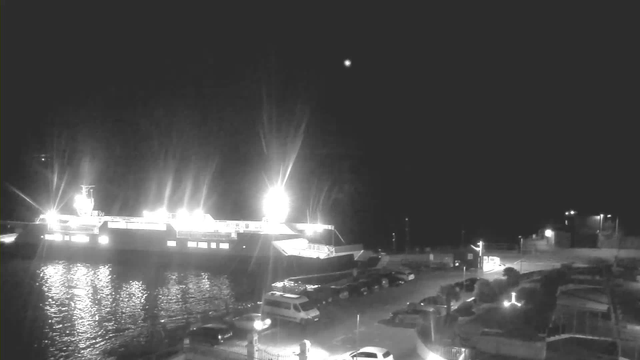 A brightly lit ferry docked at a pier during the nighttime, casting reflections on the water. Surrounding the harbor are parked vehicles, and there are faint lights from nearby buildings and additional structures in the background. A small, dim light appears in the sky above. The scene is primarily monochromatic, highlighting the contrast between the illuminated ferry and the dark surroundings.