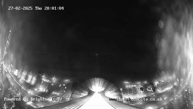 A black and white webcam image capturing a view of a cityscape at night. The bottom of the image shows a well-lit area with structured buildings, possibly featuring illuminated signs or vehicles. In the center, a bright light source appears at the horizon, suggesting a street or road leading into the distance. The upper part of the image is predominantly dark, with faint outlines of clouds or atmospheric features. The timestamp indicates it is Thursday, February 27, 2025, at 20:01:04. The watermark in the corner states "Powered by Brighton CCTV."