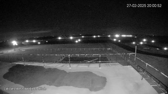 A nighttime view of an airfield with low visibility. In the foreground, there's a flat surface with a dark area on the left, possibly indicating a structure. A railing runs along the edge. In the background, several lights are visible on the airfield, suggesting the presence of runway lights. The sky is dark with minimal cloud coverage, and the timestamp indicates it is 8:00 PM on February 27, 2025.