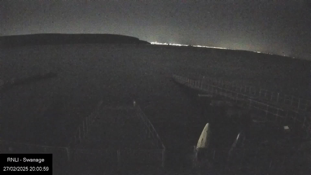 A dark scene featuring the outline of a calm body of water with distant lights twinkling on the horizon. In the foreground, there are blurred shadows of boats and a jetty extending into the water. The image is taken at night, and the overall atmosphere is tranquil but dimly lit.