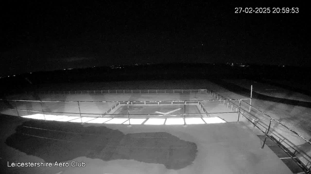 A dark nighttime scene from a webcam at Leicestershire Aero Club. In the foreground, there is a flat rooftop with a railing around it, where part of the surface is illuminated, revealing a rectangular landing area marked with a white cross. The background contains distant lights and an open sky, suggesting a rural location. The time stamp in the corner indicates the image was captured on February 27, 2025, at 20:59:53.