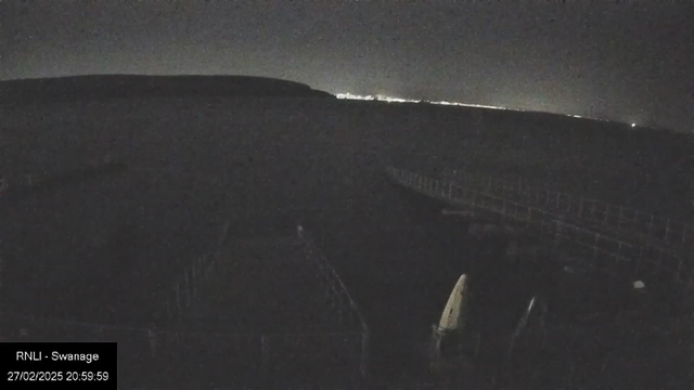 A dimly lit scene at night showing a body of water with a faint horizon line. In the foreground, there are silhouettes of structures that resemble boats or jetty sections, but details are obscured in darkness. The background features a distant line of lights, possibly from a town or city, creating a subtle glow against the night sky. The overall atmosphere is tranquil but shadowy.