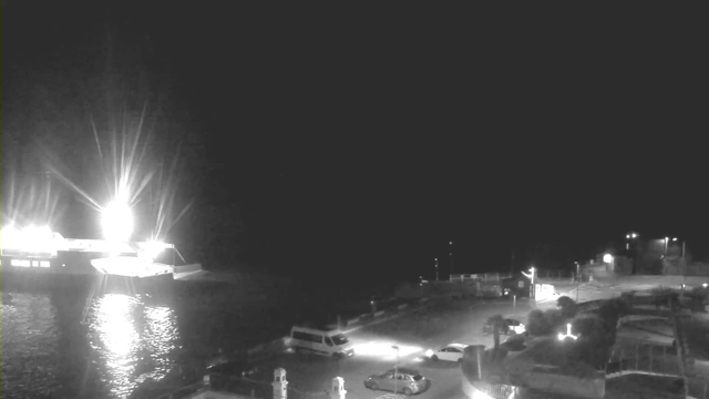 A nighttime view of a waterfront area. A bright, illuminated ferry is docked on the left side, casting reflections on the water. In the foreground, several vehicles are parked, including a van and two cars, with streetlights illuminating parts of the scene. The background is mostly dark, suggesting it is late at night, with faint outlines of land in the distance.