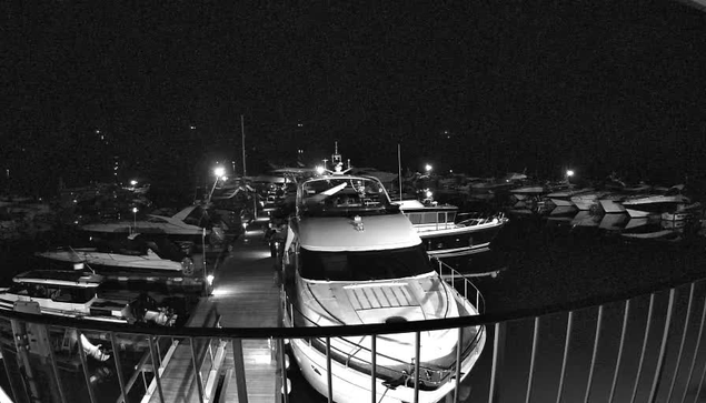 A view of a marina at night featuring several boats docked. The scene is dimly lit with lights from the boats reflecting on the water. A large yacht is prominently positioned in the foreground. Wooden walkways connect the docks, and a protective railing is visible in the lower part of the image. The background includes additional boats, some partially obscured by darkness.