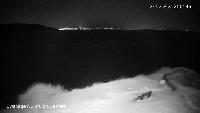 A dark scene depicting a shoreline at night with gentle waves crashing against a rocky area. In the foreground, there is a wooden bench facing the water. The background features a calm sea reflecting faint distant lights, which suggest a coastal town or city across the bay. The overall mood is tranquil and nighttime, with no visible people or activity.