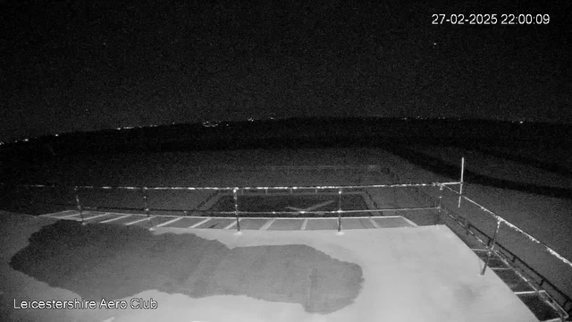 A dark landscape captured from a webcam at the Leicestershire Aero Club. The foreground features a flat surface with a railing and a shadowy patch suggesting the presence of a structure. In the background, a vast, dark sky is visible, dotted with small, faint lights indicating distant sources. The image is primarily monochromatic, emphasizing the absence of significant detail due to low light conditions. A timestamp displaying "27-02-2025 22:00:09" appears in the corner.