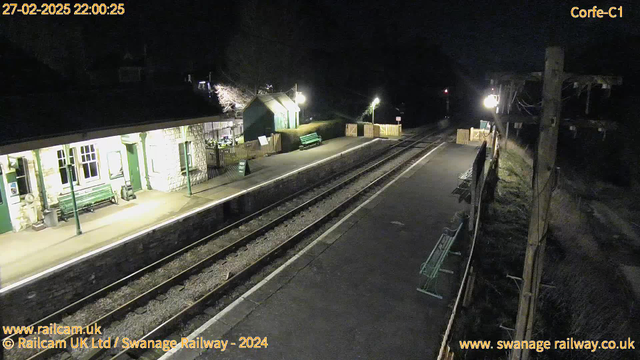 A dark scene shows a railway station platform with stone walls. There are benches along the platform, and a wooden fence in the background. One side has an illuminated area with a green building, and there are dim lights casting a glow. The railway tracks run through the center, leading into darkness. A sign indicating "WAY OUT" can be seen on the ground near the building. The atmosphere is quiet and nighttime.