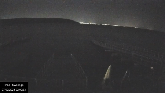 A dark scene captured at night, with a coastline visible in the distance where a faint light glimmers. In the foreground, there are outlines of structures that appear to be part of a pier or walkway. The image is mostly dark without any prominent features. The timestamp at the bottom indicates the image was taken on February 27, 2025, at 22:00:33, and a label identifies the location as RNLI - Swanage.