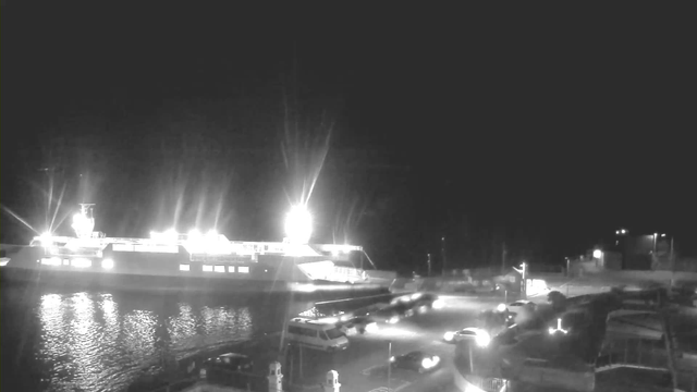 A nighttime scene at a dock with a ferry docked on the left side. The ferry is brightly lit, reflecting on the water. In the foreground, several vehicles are parked and moving near the edge of the dock. There are lights illuminating parts of the area, while shadows create a contrast against the dark background. The overall atmosphere is calm and slightly industrial, with water visible in the lower left corner.