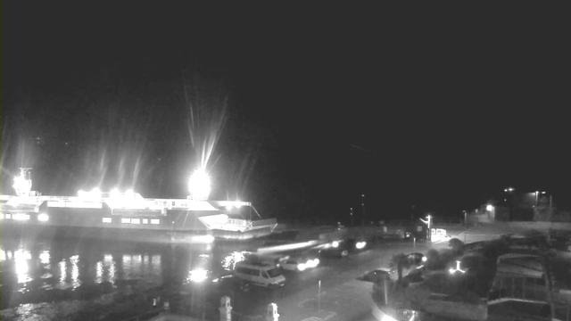 A nighttime scene at a harbor, featuring a large illuminated ferry docked on the left side of the image. The water reflects the lights from the ferry. On the right, there is an area with several parked vehicles and a roadway. Some faint lights can be seen in the background, indicating nearby structures or surroundings. The overall atmosphere is dark with bright spots from the ferry's lights.