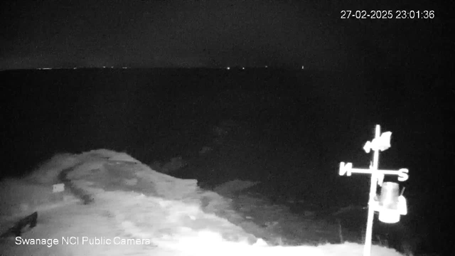 A dark scene depicting a body of water at night. The horizon is barely visible, with distant lights appearing as faint spots on the horizon. In the foreground, there is a partially obscured shoreline with waves lapping at the edge. To the right, a weather vane indicates directions, with "N" (North) and "S" (South) marked clearly. The image is monochrome, enhancing the silhouette effect of the landscape. The timestamp in the corner shows "27-02-2025 23:01:36."