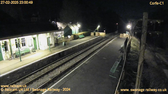 An empty railway station at night, illuminated by artificial lights. The platform features several green benches and a sign indicating an exit. A wooden fence borders the platform, and a small building with stone walls and large windows is visible on the left. Two railway tracks run along the length of the platform, which is mostly dark, with a few shadows cast from surrounding trees and structures.