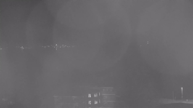 A dark, nighttime scene viewed from a webcam. The foreground features the silhouette of a building with a few lit windows, while the background shows distant lights that could be from a city or waterfront. The entire image has a hazy and foggy quality, making details less clear.