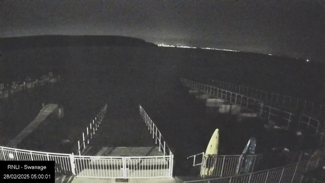 A dimly lit scene of a coastal area at night. In the foreground, a wooden walkway leads towards the water, with railings on either side. Two kayaks, one yellow and one blue, are positioned on the left side of the image, resting on the ground. The water appears dark, blending into the night sky, with faint lights visible in the distance along the horizon. The date and time are displayed at the bottom left corner of the image, indicating it is early morning on February 28, 2025.