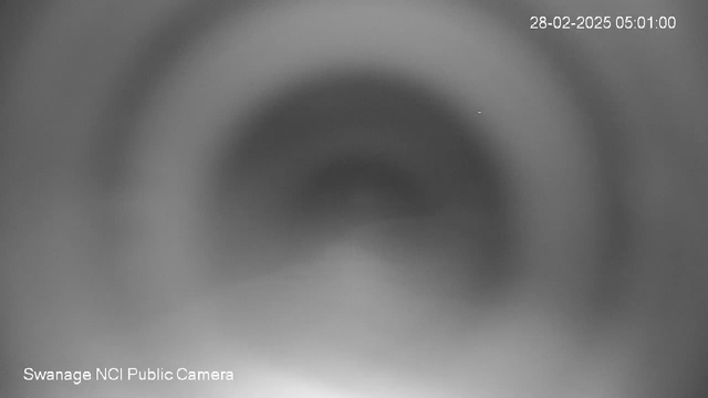 A blurred black and white image taken from a webcam, showing a circular and somewhat hazy pattern, possibly indicating a view of a lens or aperture. The background fades into lighter tones at the bottom. There is a timestamp in the top right corner indicating the date and time as 28-02-2025, 05:01:00. The bottom left corner displays "Swanage NCI Public Camera."