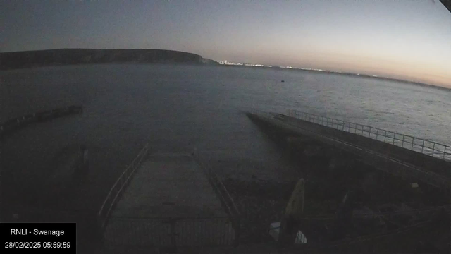 A dimly lit seaside view at dawn. The water appears calm with gentle waves, reflecting faint light from the horizon. In the foreground, a stone pier extends into the water, lined with a railing along its edge. To the left, a rocky coastline is visible, while the distant shore displays a soft glow from city lights. The sky transitions from dark above to lighter near the horizon, indicating the approach of sunrise.