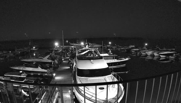 A dimly lit marina at night, featuring several boats docked closely together. The scene is monochrome, with a wooden walkway visible leading toward the boats. There are small lights illuminating parts of the marina, creating reflections on the water. The overall atmosphere is calm and quiet, with no visible people in the area.