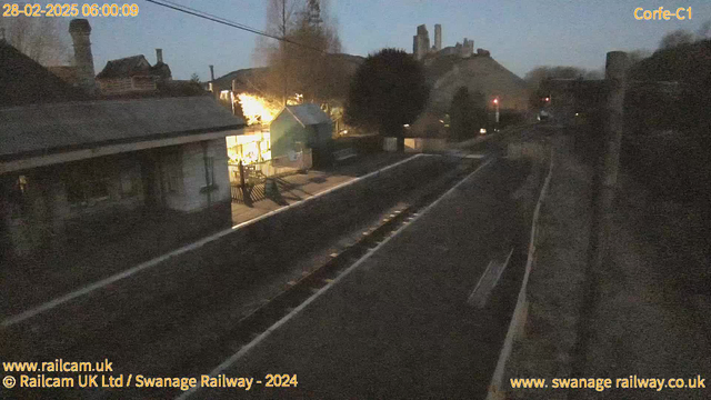 A dimly lit train station at dawn, featuring a wooden platform with faint railway tracks leading into the distance. On the left, there is a small building partially obscured by trees, and a brightly lit structure with a patio and fencing that contrasts with the surrounding darkness. In the background, there are hills and remnants of an old castle or ruins. The sky is lightening, indicating early morning, and red signal lights can be seen in the distance on the right side.