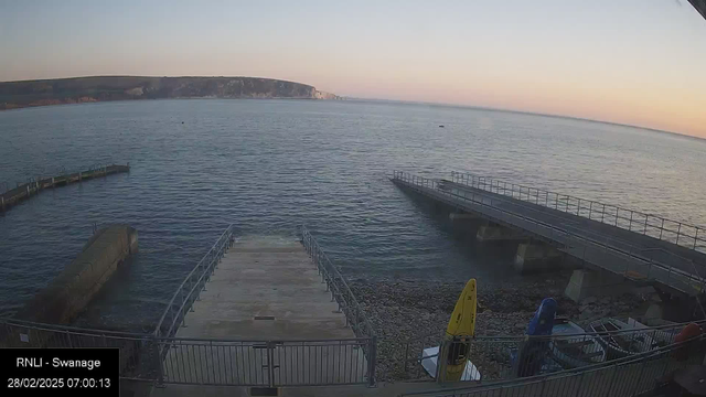 A coastal scene showing a calm body of water. In the foreground, there is a wide concrete ramp leading down to the water, bordered by metal railings. To the right, a short pier extends out into the water, with a second pier visible further back. Several boats are moored nearby, including two colorful kayaks in yellow and blue. The horizon features a distant cliffside, with the early morning sky transitioning from soft pink to light blue, indicating sunrise. The overall atmosphere is serene and tranquil.