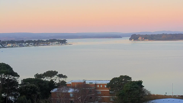 A serene view of a body of water at dawn, reflecting soft pastel hues of pink and orange in the sky. The shoreline is dotted with a mixture of buildings and trees, with some boats anchored nearby. On the left, a collection of buildings can be seen, while on the right, a distant landmass features a structure, possibly a house or a small castle, surrounded by lush greenery. The water appears calm, with gentle ripples.