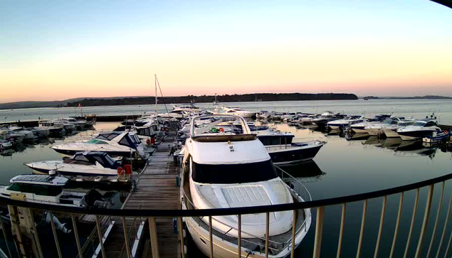 A marina at sunrise, featuring numerous boats docked along wooden piers. The water is calm and reflects the pastel colors of the sky, transitioning from soft blues to warm oranges and pinks. A large white boat is prominently positioned in the foreground, with several smaller boats lined up behind it. The scene conveys a peaceful and serene atmosphere.