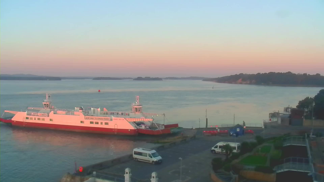 A ferry boat, painted in white with red at the bottom, is docked at a harbor. The water is calm, reflecting the soft pastel colors of the sky at dawn, including pink and orange hues. There are distant hills and small islands visible across the water. A white van is parked near the dock, and several other vehicles, including a blue structure, are also visible on the harbor's edge. Lush greenery and a landscaped area with steps lead down to the water from the right side of the image.