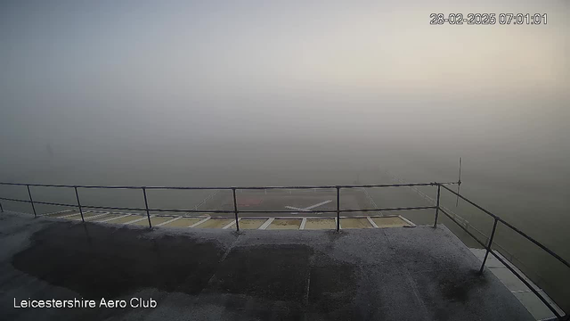 A foggy scene from a webcam shows a low visibility view from a balcony at the Leicestershire Aero Club. The foreground features a railing with a flat surface below, and there are hints of a runway or landing area barely visible through the thick mist. The lighting is soft, indicating early morning, and the overall atmosphere is muted and grey. The date and time are displayed in the top right corner, showing 28th February 2025, 07:01:01.