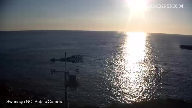 A scenic view of the ocean during sunrise. The sun is positioned in the upper right corner, casting a bright reflection on the water's surface. Gentle waves can be seen, and a weather vane with arrows indicating direction stands on the left side. The sky transitions from light blue to a warm golden hue near the horizon. The date and time are displayed in the upper corner.