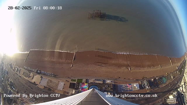 A panoramic view of a beach with calm waves lapping at the shore. The sandy beach is visible, with several wooden structures extending into the water. In the foreground, there are rooftops of buildings and colorful installations. The sky is bright, indicating daytime. A pier-like structure can be seen in the water. The image is slightly tilted, giving a sweeping perspective from an elevated position.