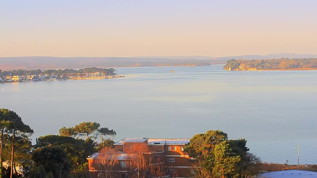 A tranquil water scene with a calm river or lake, reflecting soft daylight. In the foreground, there are trees and a building with bluish rooftops. The background features a shoreline lined with boats and buildings, with landmasses extending into the distance. The sky is clear, transitioning from soft pinks to light blues as it meets the horizon.