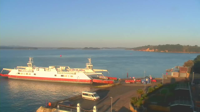 A white ferry with red trim is docked at a waterfront, with a calm blue body of water in the background. The sky is clear and bright. A white van is parked near the dock, and various structures line the shore, including a small building and some barriers. In the distance, green hills and trees are visible near the horizon.