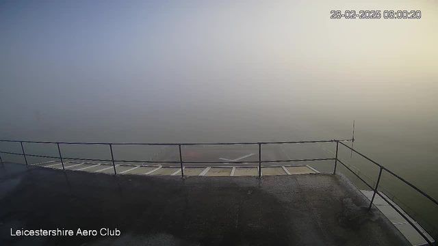 A foggy scene viewed from a rooftop, showing a railing along the edge. The visibility is very low, making the background indistinguishable. There are subtle outlines of ground structures, but they are shrouded by mist. In the lower corner, text indicates the location as "Leicestershire Aero Club," with a timestamp above indicating the date and time.