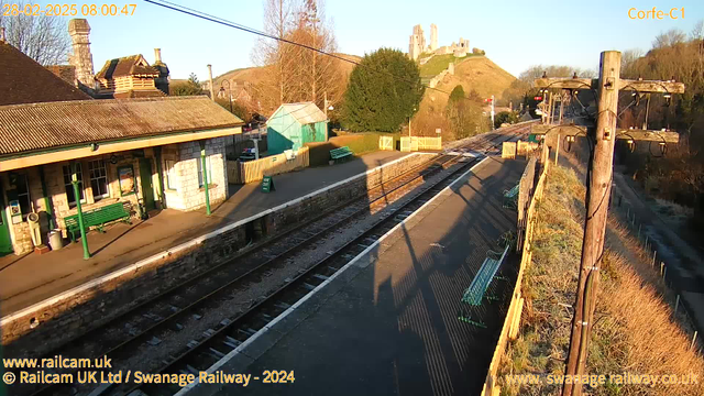 A sunlit railway station with a stone building featuring a sloped roof and green benches. The platform is flanked by railroad tracks extending into the distance. In the background, a hill rises with a castle ruin at the top, surrounded by trees. The sky is clear, indicating morning light. There are signs and a green shed nearby, with wooden fencing and a “WAY OUT” sign visible. Power lines stretch overhead.