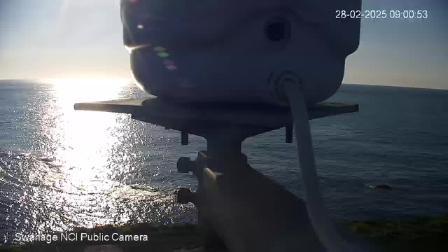 A close-up view of a webcam mounted on a pole, overlooking a tranquil ocean scene. The water reflects bright sunlight, creating a shimmering effect. The horizon is clear with a slight gradient from blue to light hues. A timestamp in the corner indicates the date and time.