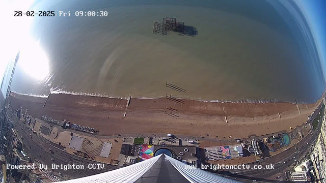 The image shows a panoramic view of a beach and the sea, captured from a high vantage point. The beach features a mix of sand and pebbles, with a few people walking along the shore. In the distance, there are visible wooden structures in the water, likely remnants of a pier. The water is calm with gentle waves, reflecting sunlight. On the beach, structures such as amusement rides and colorful tents can be seen, indicating a lively atmosphere. The sky is clear with a gradient of blue. The scene is timestamped with "28-02-2025, Fri 09:00:30" in the top left corner. The bottom of the image displays the text "Powered By Brighton CCTV" and the website of Brighton CCTV.