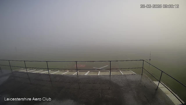 A foggy view from a raised platform at the Leicestershire Aero Club. The scene is primarily gray with limited visibility, obscuring the ground and distant objects. A railing surrounds the platform, and there are hints of structures or runway markings below, but they are difficult to see due to the dense fog. The timestamp indicates the image was taken at 9:00 AM on February 28, 2025.