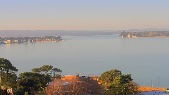 A calm body of water stretches into the distance, reflecting soft morning light. On the left, a small marina with several boats is lined along the shore, while buildings can be seen in the foreground, primarily made of brown brick. Lush green trees frame the bottom of the image, and hills are visible in the background, creating a tranquil and serene landscape.