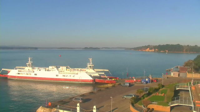 A white ferry with red accents is docked at a pier beside calm, blue water. In the background, a landscape of green trees and a sandy shoreline is visible. The sky is clear and blue, suggesting a sunny day. There are parked cars and some red barriers along the waterfront, as well as a small building with a blue structure near the pier.