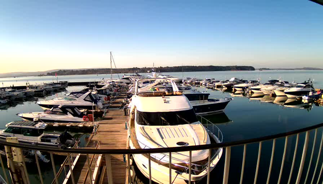 A panoramic view of a marina during daylight. Numerous boats and yachts are docked along a wooden pier, reflecting in the calm water. The background features a clear sky and distant hills, suggesting a tranquil environment. The scene conveys a sense of leisure and activity among the boats.