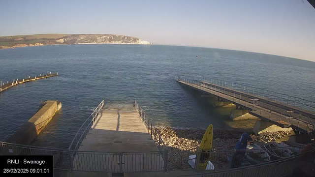 A coastal scene featuring calm blue waters with gentle waves. In the foreground, there are two piers extending into the water, one concrete and another wooden, both with railings. A sandy and rocky shore is visible along the bottom of the image, where several colorful kayaks and equipment are stored. In the background, white cliffs rise above the water under a clear blue sky.