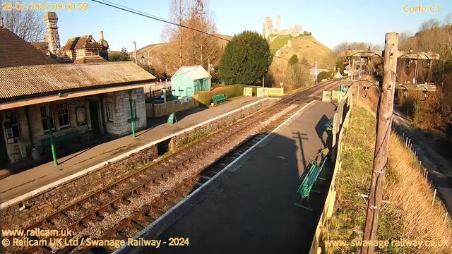 A view of a train station with a stone building on the left featuring a sloped roof and several windows. A wooden platform is visible with green benches and a small blue shed behind. There are two railway tracks running parallel with gravel in between. On the right, a wooden utility pole supports power lines. In the background, a hillside rises with a castle-like structure at the top, surrounded by trees. The sky is clear and sunny.