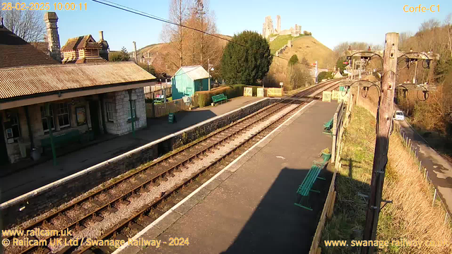 A view of a railway station platform, with wooden benches along the edge. In the background, there is a hill with ruins of a castle. The sky is clear and blue, and the sun is shining. To the left, there is a building with a sloped roof and stone walls, and a light blue shed nearby. The tracks run parallel to the platform, and there is a pole with overhead wires on the right side of the image. Green bushes and trees are visible around the area.