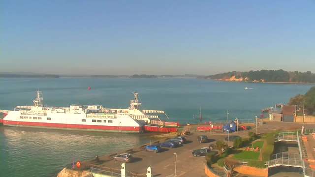 A sunny harbor scene featuring a large white and red ferry docked at a pier. The water is calm and blue, with a few boats in the distance and a red buoy nearby. There are several parked cars along the waterfront, and a pathway running beside the water. The shoreline has greenery and some buildings. The sky is clear with no visible clouds.