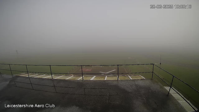 A foggy scene viewed from a balcony of an aerodrome, showing a blurred green field below. The visibility is low, making it difficult to see far into the distance. A landing strip is faintly visible with white markings, and the foreground features a railing. The sky appears gray due to the mist.