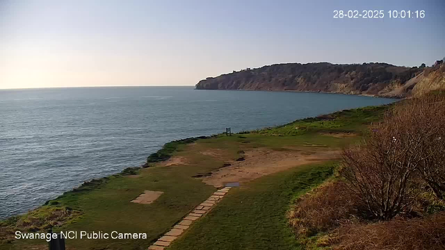 A scenic coastal view with blue ocean waters stretching out to the horizon. The shoreline features grassy areas, with a paved path leading toward the water. In the background, a rugged cliff rises, partially covered with trees. The sky is clear and sunny, indicating a bright day.