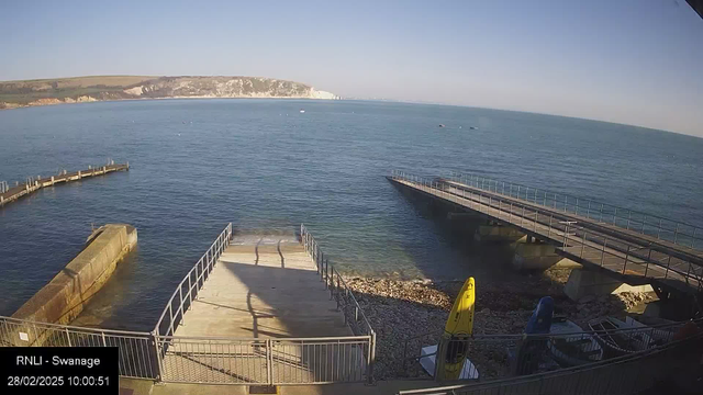 A wide view of a coastal area showing calm blue water under a clear sky. In the foreground, there are two piers extending into the water, one of which is made of concrete with a metal railing. On the right side, there are several colorful kayaks stored on the shore. A rocky beach is visible, and in the distance, a white chalk cliff rises above the waterline. The scene is bright and sunny, depicting a peaceful day at the seaside.