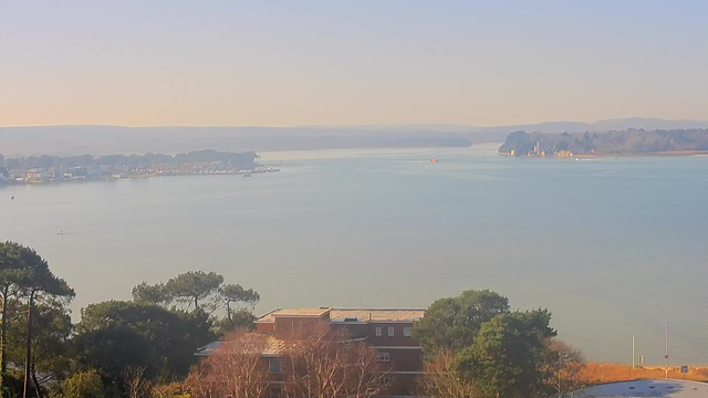 A serene view of a calm body of water stretches towards the horizon, reflecting soft colors of blue and pastel tones from the sky. In the foreground, there are patches of green trees, with a building partly visible. To the left, a line of boats is anchored along the shore. In the distance, gentle hills are outlined against the sky. A small boat can be seen moving on the water, and a hazy coastline with a few structures is visible on the right. The atmosphere appears tranquil and clear, indicating a sunny day.