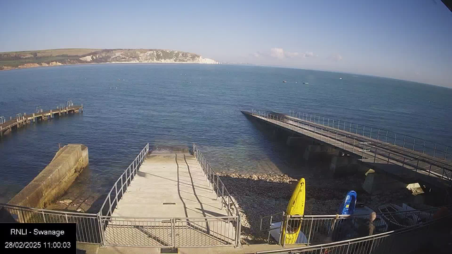 A view of a serene seaside area featuring calm waters. In the foreground, a concrete and metal ramp leads down to the water, bordered by a railing. To the right, there are two boats, one yellow and one blue, resting on the shore. In the background, a wooden pier extends into the water on the left side. The distant shore includes white cliffs and greenery under a blue sky with a few clouds.